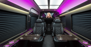 Luxury jet sprinter van interior featuring black diamond-stitched leather captain's chairs, dark wood folding tables, and vibrant purple LED accent lighting. The cabin includes a large ceiling light panel, privacy window shades, and a flat-screen TV at the front.
